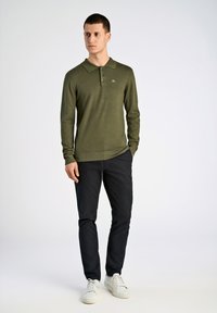 Olivgrünes Langarm-Polo mit Kragen und einer Knopfleiste mit drei Knöpfen, kombiniert mit schwarzen Hosen und weißen Sneakers. Weiche Stricktextur.