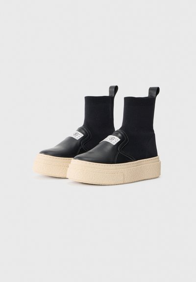 Svarta läderplattformssneakers med en texturerad beige sula och en stickad sock-liknande överdel. Har en dragflik och varumärkets logotyp på sidan.