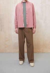 Helles rosa Hemd über einem grün-blauen gestreiften T-Shirt, kombiniert mit braunen weit geschnittenen Hosen und cremefarbenen Schuhen vor einem neutralen Hintergrund.