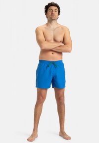 Calções de banho azuis feitos de tecido leve, com cós elástico, cordão verde e corte confortável.