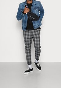 Chaqueta de mezclilla sobre una camiseta negra, combinada con pantalones de cuadros grises y zapatillas negras. Se lleva una riñonera negra cruzada sobre el torso.