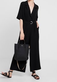 Combinaison noire à manches courtes, taille ceinturée et décolleté en V profond. Assortie à un sac fourre-tout noir et des sandales à lanières noires.