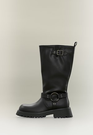 Botte en cuir noire mi-mollet avec une semelle épaisse, une sangle décorative à boucle près de la cheville, et une sangle réglable avec boucle métallique près du haut.