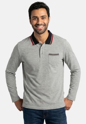 Hombre sonriendo que lleva una camisa polo gris claro de manga larga con cuello negro y detalles en rojo y blanco, y vaqueros azules.