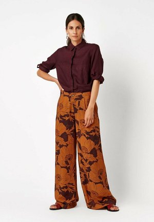 Frau steht mit der Hand auf der Hüfte, trägt ein bordeauxfarbenes Langarmshirt und weit geschnittene Hose mit großem orangefarbenem Blumenmuster und Sandalen.