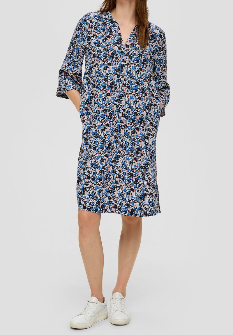 Femme portant une robe fleurie ample mi-longue en bleu, rose et noir avec des manches trois-quarts et des baskets blanches, les mains dans les poches.