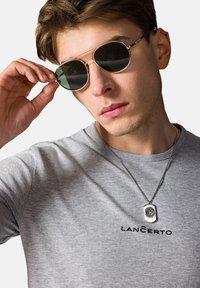 Okulary przeciwsłoneczne w złotej oprawie z ciemnymi soczewkami, szary t-shirt z logo "LANČERTO", srebrny naszyjnik z prostokątnym wisiorkiem.