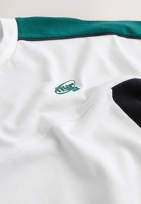 Camisa blanca con panel verde en el hombro, detalle negro en la manga y pequeño logo verde en el pecho sobre fondo de tela lisa.