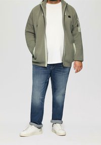 Zelená zipová mikina s bočními kapsami a světle zelenými detaily, oblečená přes bílé tričko a modré džíny. Bílé tenisky dokončují outfit.