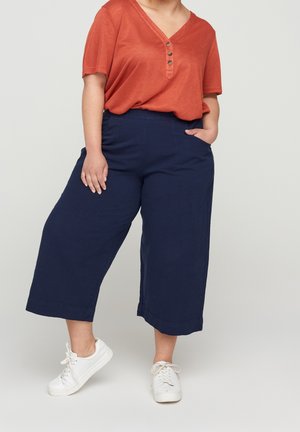 Personne portant un haut orange ample à manches courtes, un pantalon large bleu marine court et des baskets blanches, debout avec une main dans la poche.