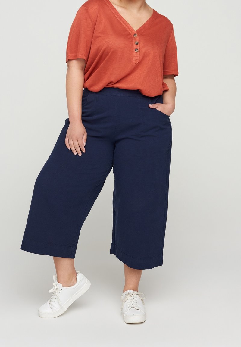 Personne portant un haut orange ample à manches courtes, un pantalon large bleu marine court et des baskets blanches, debout avec une main dans la poche.