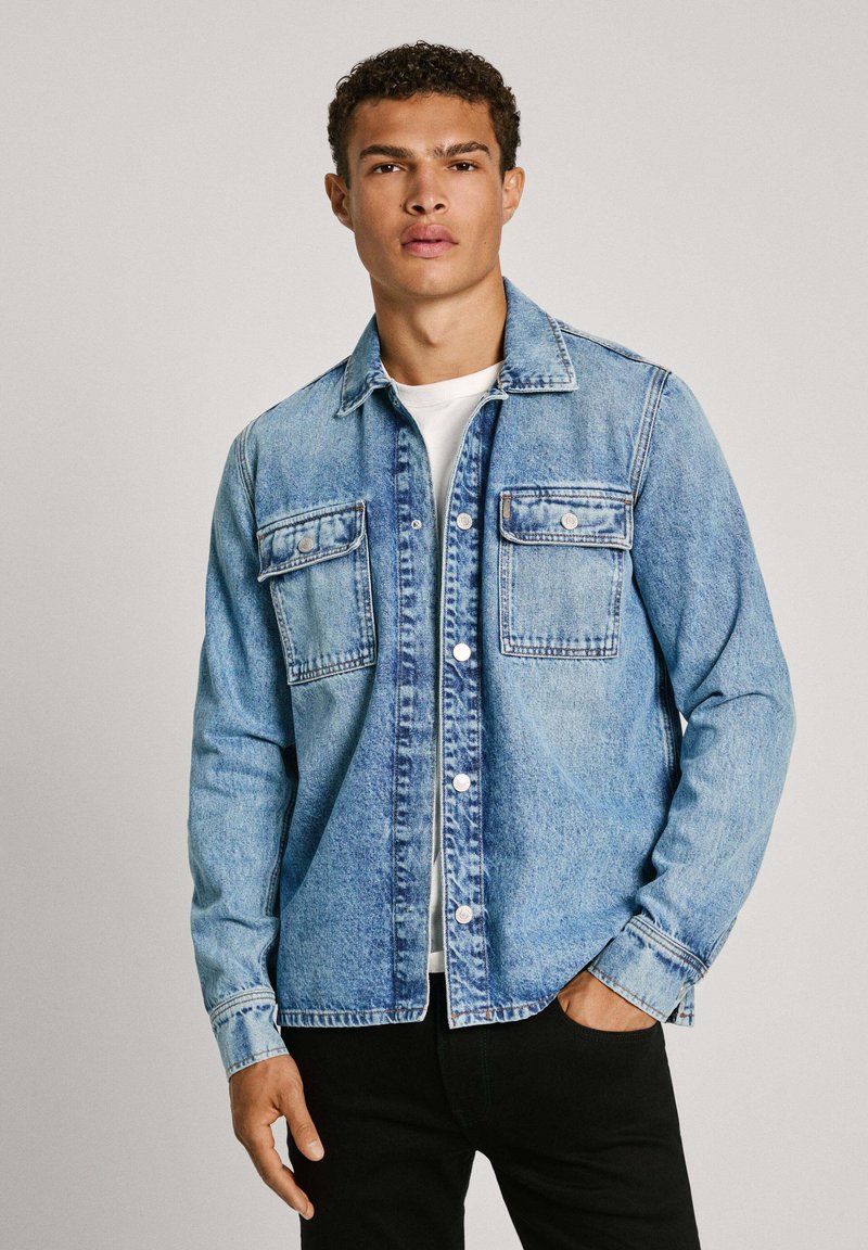Joven con cabello rizado vestido con una chaqueta de mezclilla azul claro sobre una camisa blanca y pantalones negros, posando frente a un fondo liso.