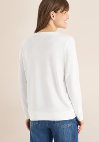Haut à manches longues blanc avec encolure ronde, tissu lisse et poignets côtelés, associé à un jean bleu. Design épuré et minimaliste sans motifs.