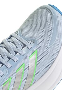 Zapatilla deportiva azul claro con parte superior de malla, detalles en plata y verde, superficie texturizada y una suela blanca acolchada. Incluye cordones.