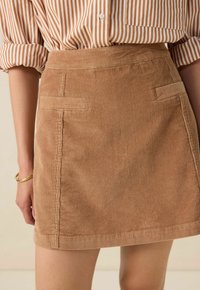 Mini jupe en velours côtelé marron avec des rayures verticales, dotée de deux poches avant et d'une taille lisse. Texture lisse et forme structurée.