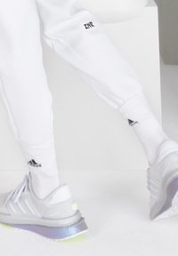 Białe spodnie sportowe z nadrukiem "ZNZ", w po łączeniu z szarymi butami sportowymi z odblaskową podeszwą i czarnym logo Adidas. Widoczne białe skarpetki.