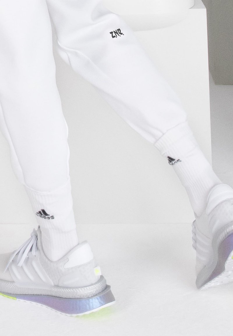 Białe spodnie sportowe z nadrukiem "ZNZ", w połączeniu z szarymi butami sportowymi z odblaskową podeszwą i czarnym logo Adidas. Widoczne białe skarpetki.