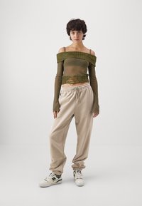 Durchsichtige olivgrüne Off-Shoulder-Bluse mit gerippten Akzenten, kombiniert mit lockeren beigen Jogginghosen und weißen Sneakers mit grünen Details.
