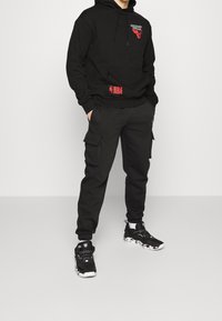 Felpa con cappuccio nera con logo dei Chicago Bulls e patch rossa NBA, abbinata a pantaloni cargo neri e sneakers a fantasia bianca e nera.