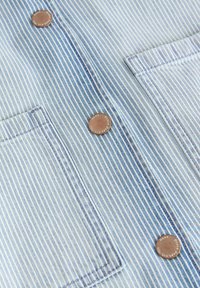 Tela de denim azul clara con rayas verticales blancas. Cuenta con dos bolsillos en el pecho con botones de cobre y detalles de costura en contraste.