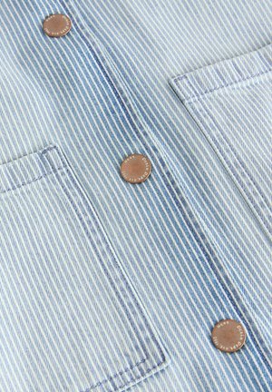 Tissu en denim bleu clair avec des rayures blanches verticales. Il présente deux poches sur la poitrine avec des boutons en cuivre et des détails de couture contrastés.