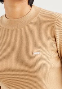 Zbliżenie na osobę ubraną w beżowy sweter z prążkowanego dzianiny z małym białym logo Levi's na piersi.