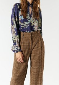 Blouse légère bleu marine à motif floral, dotée d'une encolure fendue et de poignets à volants, associée à un pantalon large à carreaux marron avec une finition texturée.