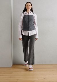 Modell som har på seg en grå denimvest over en hvit skjorte, parret med grå vide jeans og hvite platå-sandaler. Enkelt design og strukturert passform.
