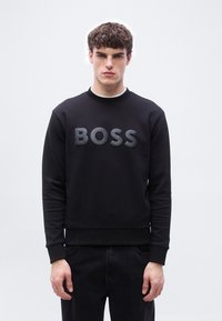 Νεαρός άνδρας με κοντά σγουρά μαλλιά που φοράει μαύρο φούτερ με το ανάγλυφο γράμμα "BOSS", στέκεται μπροστά σε απλό φωτεινό φόντο.