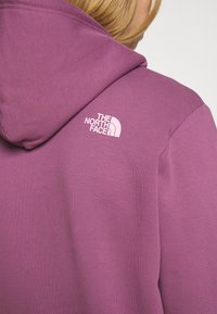 Lila hoodie med känguruficka, ribbade ärmslut och nederkant. Har en vit logotyp på övre vänstra delen av ryggen. Slät tygtextur.