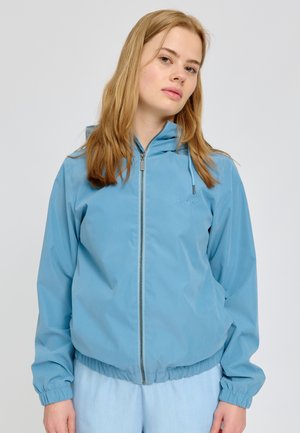 Junge Frau mit hellbraunen Haaren, die eine hellblaue Kapuzenjacke mit Reißverschluss und passende Hosen vor einem einfarbigen Hintergrund trägt.