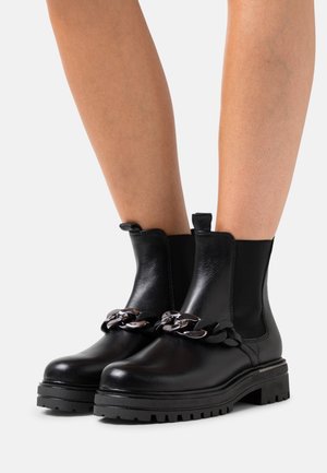 Botas Chelsea negras con suela gruesa, con un detalle de cadena plateada en la parte delantera y una lengüeta en la parte trasera para facilitar el calzado.