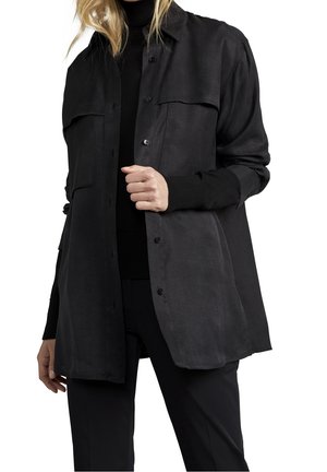 Nainen, joka pukeutuu auki napitettuun pitkään mustaan paitaan mustan pooloneuleen ja mustien housujen päällä, vaaleat hiukset löysänä.