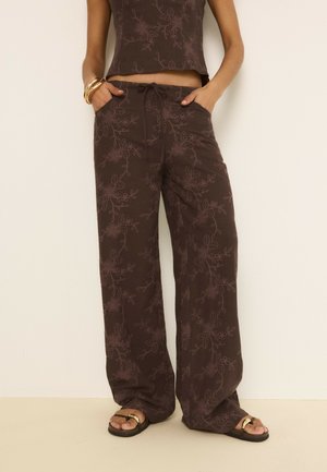 Femme portant un pantalon large marron à fleurs et un haut sans manches assorti, avec des bracelets dorés et des sandales noires avec des détails dorés.