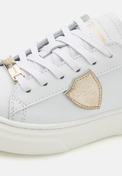 Sneaker in pelle bianca con lacci bianchi, dettagli argentati e logo a scudo color taupe. Presenta una suola in gomma bianca testurizzata e dettagli cuciti.