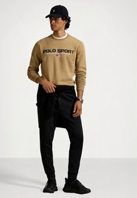 Μπεζ φούτερ με το λογότυπο "POLO SPORT", μαύρο παντελόνι και μαύρα αθλητικά παπούτσια. Το μοντέλο φοράει επίσης μαύρο καπέλο και κρατάει ένα φούτερ δεμένο γύρω από τη μέση.