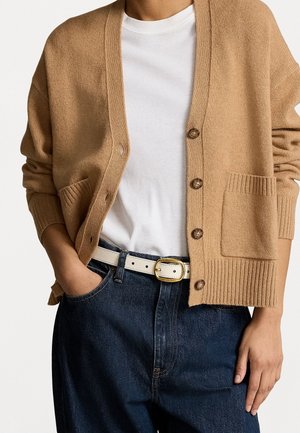 Personne portant un t-shirt blanc sous un cardigan marron boutonné avec poches avant, un jean bleu foncé et une ceinture blanche avec une boucle dorée.