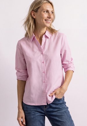 Femme portant une chemise rose clair boutonnée avec les manches retroussées et un jean bleu, souriante et regardant sur le côté avec une main dans la poche.