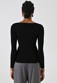 Top lungo a maniche nere a coste con scollatura quadrata, design aderente e tessuto testurizzato. Indossato con pantaloni grigi a vita alta.