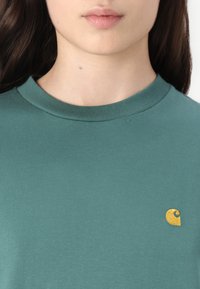 Sünnipäeva smaragdroheline puuvillane T-särk, millel on ümmargune kaelus ja vasakul väike kuldne brodeeritud logo. Sile tekstuur, keha lähedalt istuv disain.
