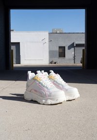 Άσπρα chunky sneakers με παστέλ ροζ, πράσινες και κίτρινες λεπτομέρειες τοποθετημένα σε μπετόν κάτω από την σκιερή είσοδο ενός βιομηχανικού κτηρίου.