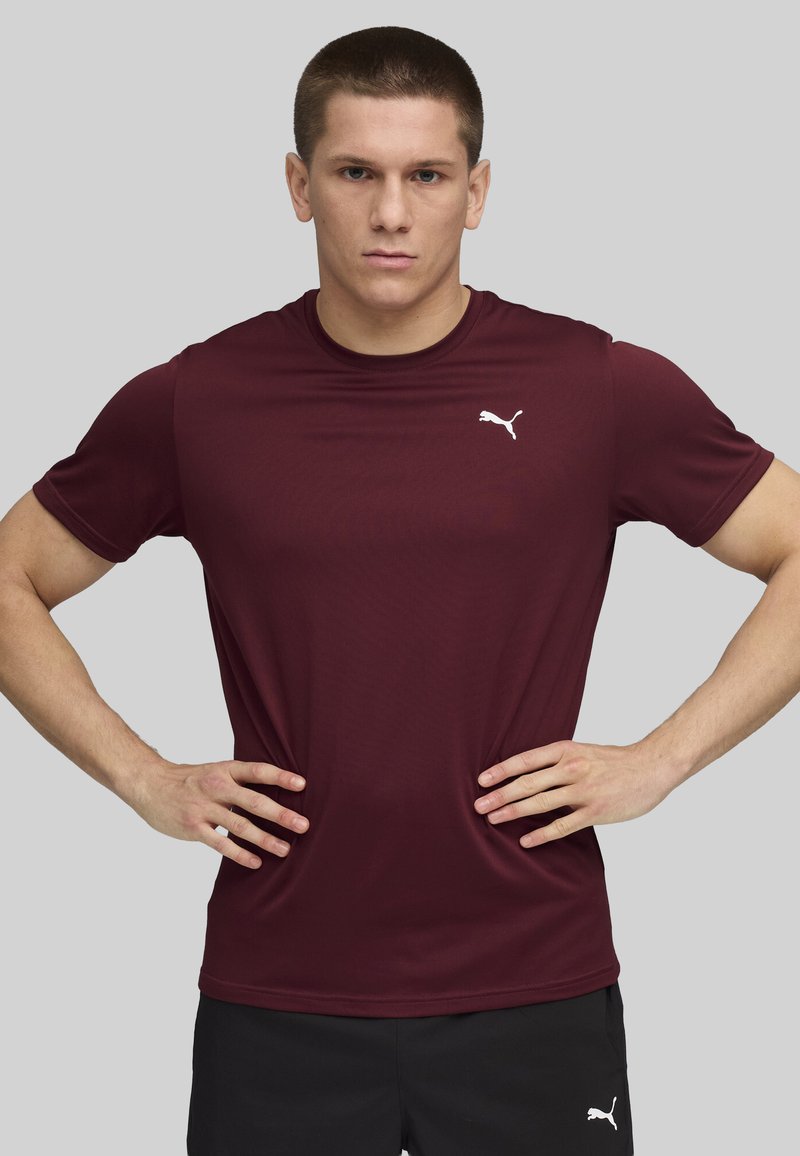 Camiseta deportiva burdeos de tejido suave, con un logo blanco de Puma en el lado izquierdo del pecho, cuello redondo y mangas cortas.
