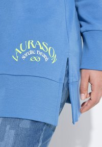 Felpa azzurro chiaro con texture a coste, fessure laterali e logo giallo neon con la scritta "LAURASON Nordic Brand" sul davanti.