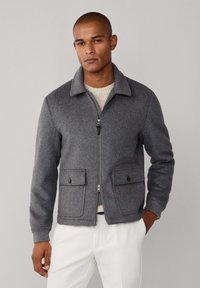 Chaqueta gris de lana con cuello, cierre frontal de cremallera, dos bolsillos con solapa y botón, y puños acanalados, usada sobre un suéter beige.