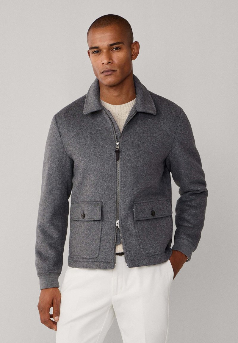 Chaqueta gris de lana con cuello, cierre frontal de cremallera, dos bolsillos con solapa y botón, y puños acanalados, usada sobre un suéter beige.