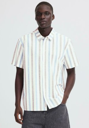 Młody mężczyzna w koszuli na guziki z krótkim rękawem w pionowe pasy w pastelowym niebieskim, beżowym i białym kolorze oraz w ciemnoszarych spodniach, stojący na białym tle.