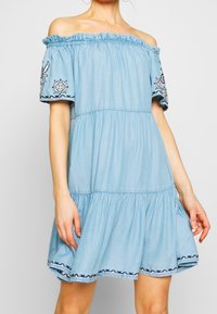 Vaaleansininen olkapäältä paljastava mekko, joka on valmistettu denimpölystä, ja siinä on elastinen kaula-aukko sekä kirjailtua lyhyet hihat, joissa on koristeellisia kuvioita.