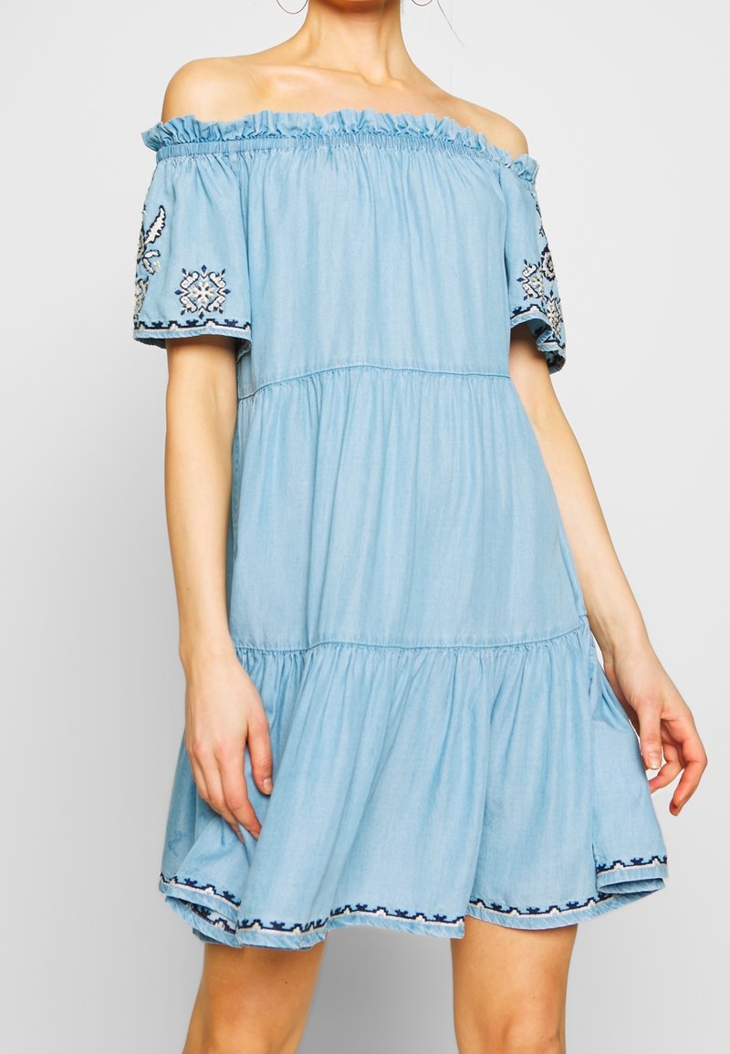 Vaaleansininen olkapäältä paljastava mekko, joka on valmistettu denimpölystä, ja siinä on elastinen kaula-aukko sekä kirjailtua lyhyet hihat, joissa on koristeellisia kuvioita.