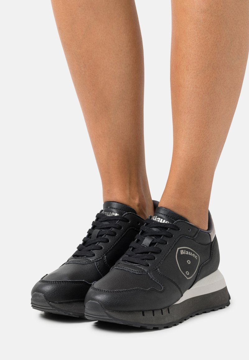 Person med svarta Blauer lädersneakers med svarta snörningar och vita detaljer på sulorna, stående mot en vit bakgrund.