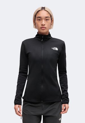 Person med kort blondt hår iført en sort lynlåstrøje og bukser med The North Face-logo, stående mod en ensfarvet baggrund.